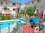 Outdoor amenity area including in ground pool with safety fencing and a small playground area, surrounded by three-story stucco apartment buildings